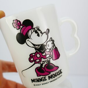 May include: White ceramic Minnie Mouse mug featuring a heart-shaped handle. The mug showcases a smiling Minnie Mouse in a pink dress and bow, holding a purse. The words "MINNIE MOUSE" and "WALT DISNEY PRODUCTIONS" are printed on the mug.