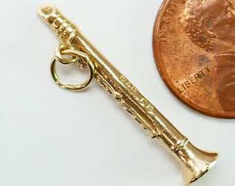 May include: A gold-colored clarinet charm with a ring for attaching to a necklace or bracelet. The charm is about the size of a US penny.