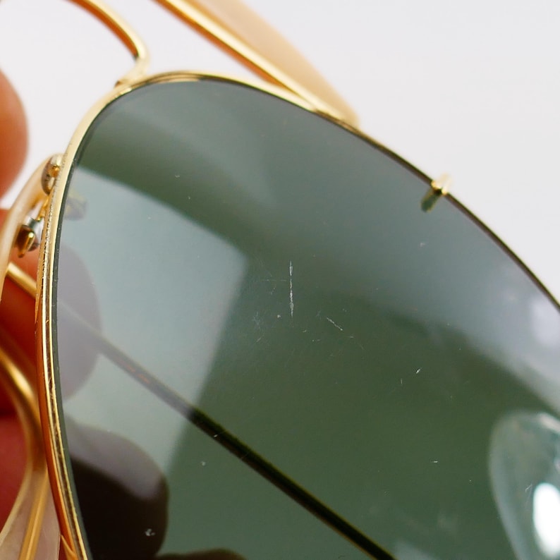 May include: Close-up of gold-framed sunglasses with green lenses. The sunglasses have a classic aviator design. The lenses show minor scratches. The gold frames are bright and shiny.