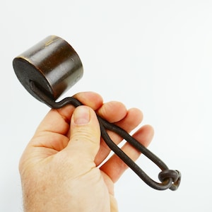 May include: A vintage brass weight with a black metal handle. The weight is round and has a smooth surface. The handle is bent into a loop at the end.