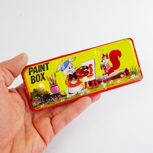 May include: A rectangular yellow paint box with a red border. The box features a cartoon illustration of a rabbit artist with a palette and easel, and a squirrel. The words "PAINT BOX" are printed on the box.