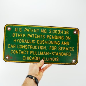 Può includere: Segno di metallo verde con testo giallo. Il segno recita "U.S. PATENT NO. 3,003,436 OTHER PATENTS PENDING ON HYDRAULIC CUSHIONING AND CAR CONSTRUCTION. FOR SERVICE CONTACT PULLMAN-STANDARD CHICAGO, ILLINOIS"