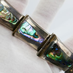 Peut inclure: Gros plan sur un bracelet en argent avec des maillons rectangulaires. Chaque maillon présente une incrustation irisée en coquille d'ormeau avec des nuances de vert, de bleu et de violet. Le bracelet est présenté sur un fond blanc.