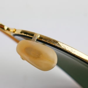 May include: Close-up of gold-rimmed sunglasses with a light-colored nose pad. The lenses are a dark green color. The gold frame shows signs of wear. The sunglasses are set against a white background.
