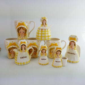 Puede incluir: Colección de utensilios de cocina de cerámica con un diseño de niña encantadora. El juego incluye tazas, una jarra de crema, un azucarero, saleros y pimenteros, y un servilletero. Las piezas son blancas con detalles amarillos y un estampado de cuadros.