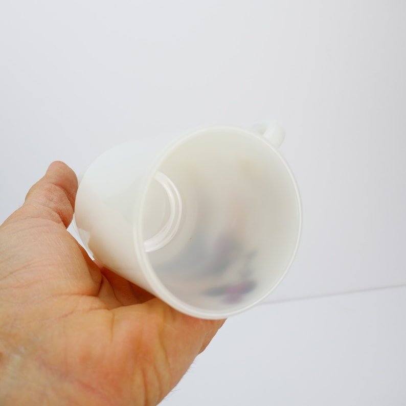 May include: A white ceramic mug with a handle, held in a person's hand. The mug has a smooth, matte finish and a simple, cylindrical shape. The interior of the mug is visible, with a hint of a design at the bottom. The mug is likely for beverages.