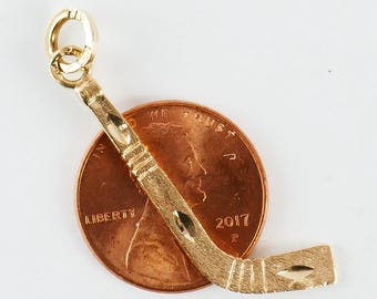 Könnte beinhalten: Ein goldener Hockey-Schlüsselanhänger, gezeigt neben einem US-Cent zum Größenvergleich. Der Anhänger ist mit einer strukturierten Oberfläche und einer Schlaufe zum Anbringen an einer Kette verziert.