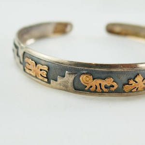 Puede incluir: Brazalete de plata con detalles dorados. El brazalete presenta una banda gris oscuro con diseños geométricos y animales dorados. El diseño de brazalete abierto permite un fácil uso. El brazalete es una pieza de joyería única.