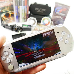 Puede incluir: Una consola de juegos portátil Sony PSP plateada que muestra la pantalla de título del juego Need for Speed Carbon Own the City. El dispositivo cuenta con una cruceta, cuatro botones de acción, un joystick y varios botones de control.