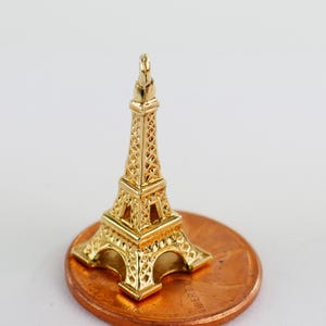 Puede incluir: Una pequeña réplica dorada de la Torre Eiffel. La torre en miniatura es muy detallada, con intrincados trabajos de celosía y una parte superior puntiaguda. Se coloca sobre una moneda de un centavo para mostrar su tamaño. El fondo es de un color suave y neutro.