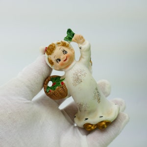 Puede incluir: Una figurita de porcelana blanca de un ángel con una cesta de acebo y una hoja verde en la mano. Lleva un vestido blanco con detalles dorados y tiene una cara sonriente.