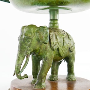 May include: A green, antique-style decorative bowl with an elephant base. The elephant is detailed with carved features and stands on a round, wooden base. The bowl has a wide, shallow design, perfect for displaying items.