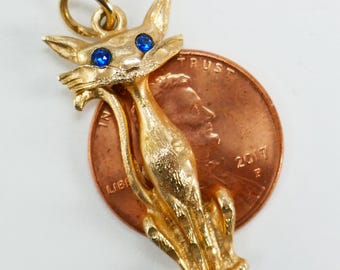 May include: A gold-toned cat charm with blue rhinestone eyes. The charm is sitting on top of a US penny for size comparison.
