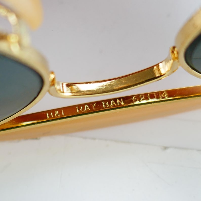 May include: Close-up of gold-framed sunglasses with dark lenses. The text "RAY BAN" is visible on the temple arm, along with the numbers "62 14". The sunglasses have a classic design.