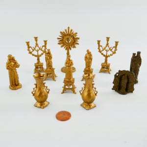 Puede incluir: Una colección de figuritas religiosas doradas, que incluyen una estatua de la Virgen María, una estatua de un santo y una estatua de un sacerdote. También hay dos candelabros dorados, un jarrón dorado y un sol dorado.
