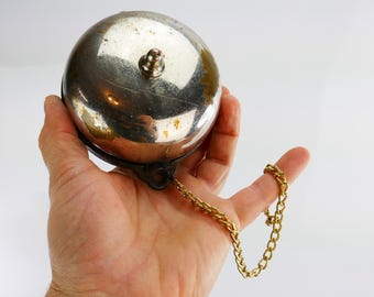 May include: A vintage silver bell with a tarnished finish. The spherical bell has a button on top and a gold-coloured chain. The bell is held in a hand against a white backdrop.