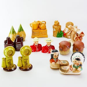 Könnte beinhalten: Eine Sammlung von vintage Keramik Salz- und Pfefferstreuern mit verschiedenen kanadischen Motiven. Die Streuer zeigen einen Mountie, ein Telefon, eine Blockhütte, eine Teekanne, ein Eichhörnchen, ein Schwein und einen indigenen Menschen, der Trommel spielt.