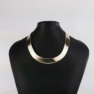 May include: A gold-coloured collar necklace displayed on a black mannequin. The necklace has a smooth, polished surface and a curved, open design. The background is a plain white.
