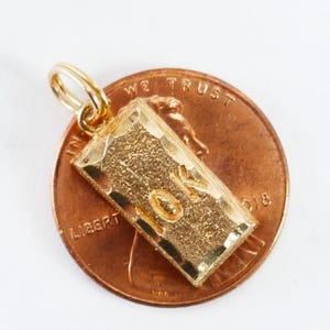 May include: A gold-colored charm shaped like a gold bar with the inscription "10K" on it. The charm is sitting on top of a US penny.