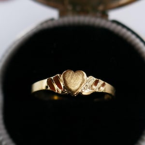 Könnte beinhalten: Ein goldener Ring mit herzförmigem Mittelteil und dekorativen Seitendetails. Der Ring ist vor einem dunklen Hintergrund platziert, der seine polierte Oberfläche hervorhebt. Ein klassisches Schmuckstück.
