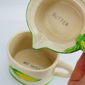 Puede incluir: Taza y tapa medidora de cerámica color crema, con ribete verde e ilustraciones de maíz. La taza tiene la inscripción "HOT WATER" en el interior, y la tapa "BUTTER". Se ve un asa en la taza.