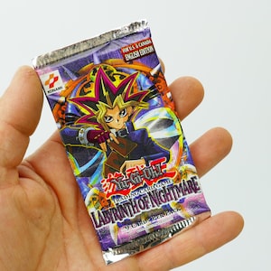 May include: A sealed Yu-Gi-Oh! Trading Card Game pack, "Labyrinth of Nightmare" English Edition, held in a hand. The pack features a character illustration with vibrant colours and the Konami logo. The text "9 Cards Per Pack" is visible.