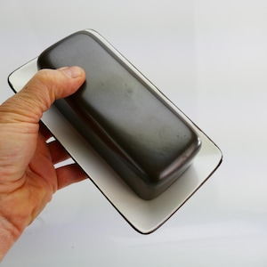 May include: A dark brown butter dish with a lid, on a white rectangular plate with a black trim. The dish is held by a hand. The dish and plate are rectangular.