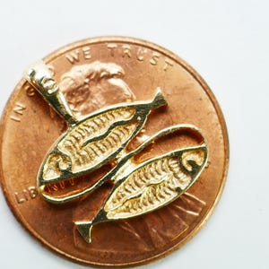 May include: Gold-toned pendant charm featuring two fish with detailed scales. The charm is shown next to a US penny for size comparison.