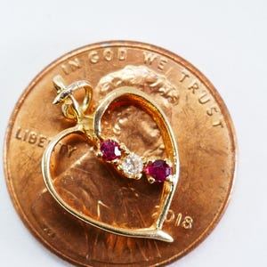 Può includere: Ciondolo a forma di cuore in oro con tre pietre preziose rosse e una sola pietra preziosa trasparente. Il ciondolo è mostrato su un penny americano per il confronto delle dimensioni.
