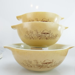 Könnte beinhalten: Ein Satz von drei ineinandergreifenden Schüsseln in einem warmen Gelbton. Jede Schüssel zeigt eine braune Illustration von Pilzen und Blättern. Die Schüsseln haben ein klassisches Design mit seitlichen Griffen.