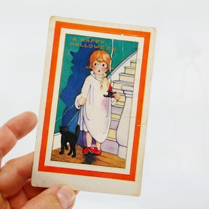 Puede incluir: Postal vintage de Halloween con un niño en camisón blanco y zapatos rojos, sosteniendo una vela. Hay un gato negro. La tarjeta tiene un borde naranja y el texto "A Happy Halloween".