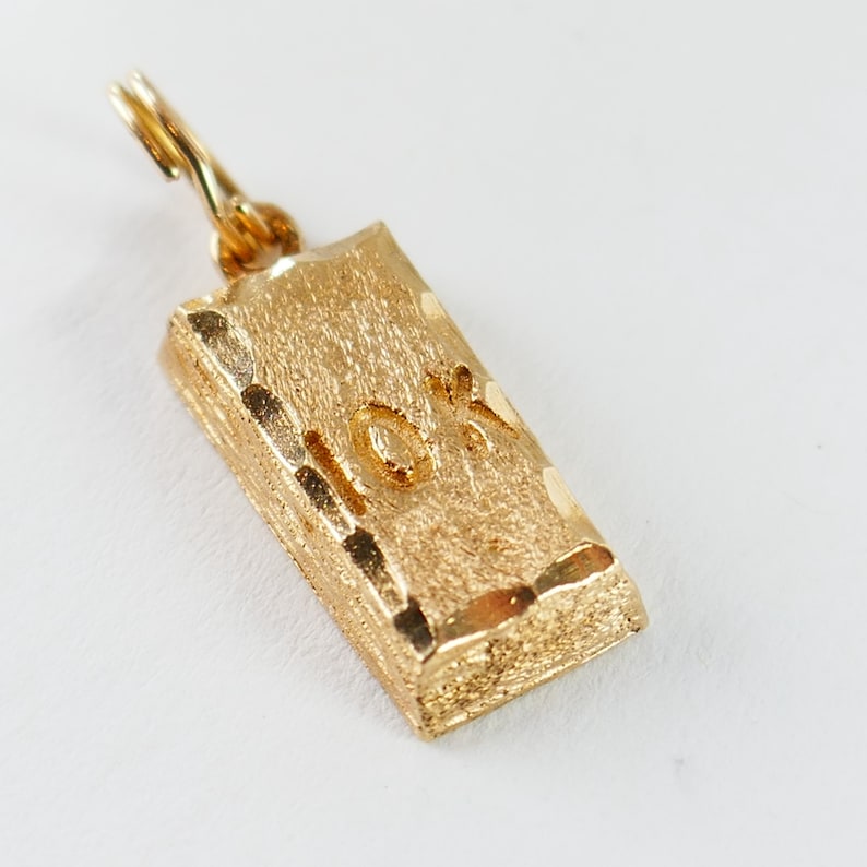 May include: Gold-colored charm shaped like a gold bar with a textured surface and the inscription "10K" on the front.