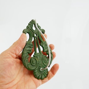 Puede incluir: Un llamador de puerta verde y ornamentado con un diseño decorativo en espiral. El llamador tiene un asa curva de color plateado. El llamador se sostiene en una mano sobre un fondo blanco. El llamador es de metal.