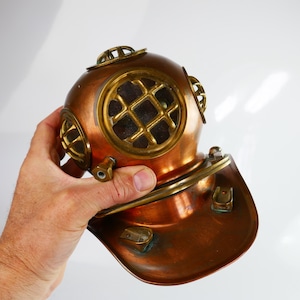 Puede incluir: Un casco de buceo vintage de cobre con detalles de latón. El casco tiene una parte superior redondeada con múltiples portillos y una base acampanada. Los portillos tienen un patrón de rejilla. El casco es sostenido por una mano.