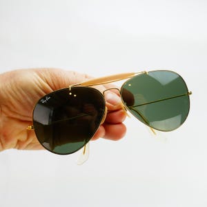 May include: Gold-framed aviator sunglasses with green lenses. The sunglasses have a double bridge and a light-colored brow bar. The Ray-Ban logo is visible on the left lens. The sunglasses are held against a white background.
