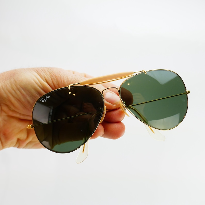 May include: Gold-framed aviator sunglasses with green lenses. The sunglasses have a double bridge and a light-colored brow bar. The Ray-Ban logo is visible on the left lens. The sunglasses are held against a white background.
