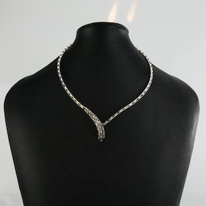 May include: A silver necklace with a textured, woven pattern. The necklace is presented on a black mannequin, highlighting its elegant, curved form. The pendant has a cluster of vertical bars.