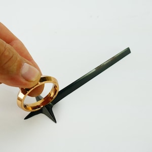 Pode incluir: Um anel de metal dourado é colocado em cima de uma flecha de metal preto com uma ponta pontiaguda.