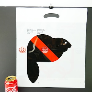 Könnte beinhalten: Weiße Plastiktüte mit einem schwarzen und roten Biber-Motiv und dem Text "Jeux de la XXle Olympiade Montreal 1976" und "Games of the XXI Olympiad Montreal 1976".