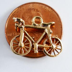 Può includere: Un piccolo ciondolo a forma di bicicletta color oro con raggi e manubrio dettagliati. Il ciondolo è posto su un penny, con le parole "LIBERTY" e "IN GOD WE TRUST" visibili. Il ciondolo è una miniatura di una bicicletta.