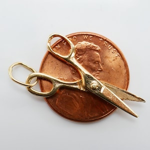 May include: A gold-coloured scissor charm with a loop for attaching to a necklace or bracelet. The charm is shown on top of a US penny.