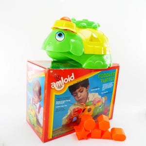 May include: A green and yellow plastic toddler turtle toy with a matching box. The turtle has a yellow hat and a colorful spin top. The box features the text "TODDLER TURTLE" and "amloid". Orange geometric shapes are in front of the box.
