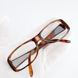 Peut inclure: Une paire de lunettes de vue en &eacute;caille de tortue brune avec des verres transparents. Les lunettes sont pos&eacute;es sur une surface blanche.