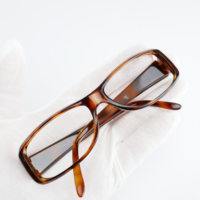 Peut inclure: Une paire de lunettes de vue en &eacute;caille de tortue brune avec des verres transparents. Les lunettes sont pos&eacute;es sur une surface blanche.