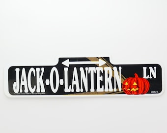 Könnte beinhalten: Ein schwarz-weißes Straßenschild mit dem Text "JACK-O-LANTERN LN" in weißen Buchstaben. Das Schild hat einen schwarzen Hintergrund mit einem goldenen und braunen Bild einer Straßenszene. Eine rote und orangefarbene Kürbislaterne befindet sich auf der rechten Seite des Schildes.