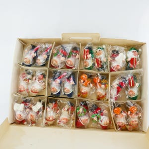 May include: A box containing 18 small, colourful, felt Christmas gnome decorations. The gnomes are wearing a variety of hats and outfits, including a red and white Santa hat, a green hat with a white pom-pom, and a blue hat with a white pom-pom.