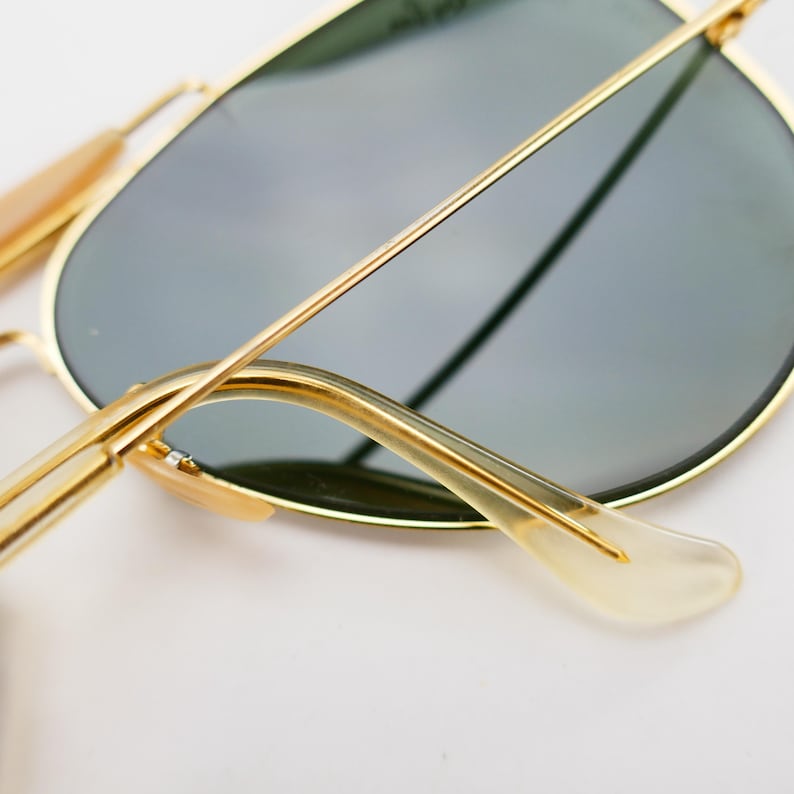 May include: Gold-framed aviator sunglasses with dark lenses. The sunglasses have a classic design with a thin gold frame and a curved brow bar. The temples are a light tan color and curve around the ears.