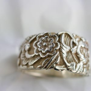 May include: A silver ring with a floral design. The ring features detailed carvings of flowers and leaves, creating a textured surface. The ring's band is wide, and the overall style is ornate and vintage-inspired.