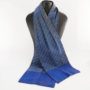May include: A blue scarf with a repeating gold and blue pattern. The scarf is draped over a black mannequin. The ends of the scarf are a solid, darker blue. The scarf is made of a soft, flowing material.