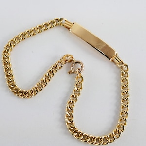 May include: A gold-colored curb chain bracelet with a rectangular plate. The bracelet features a lobster clasp and is likely made of gold-plated metal. The chain links are closely linked together.